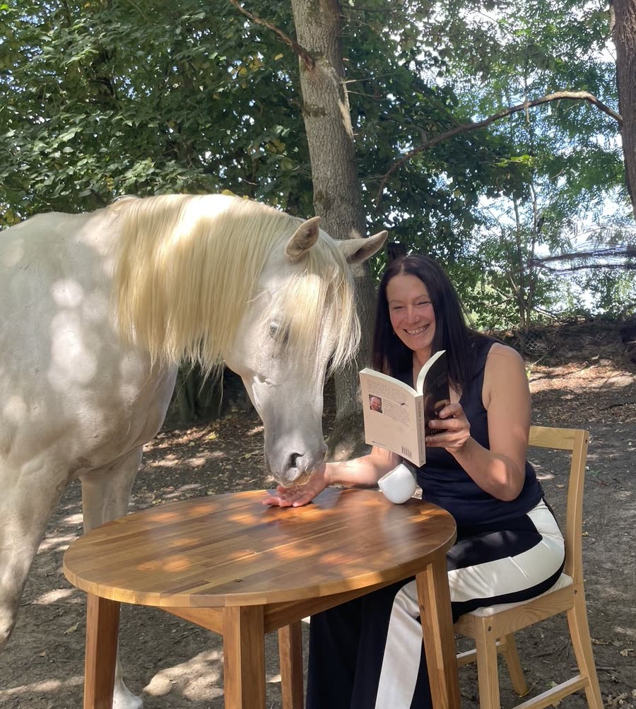 Nele Van Steertegem leest haar boek aan een tafeltje in de tuin, met een van haar paarden vlak naast zich